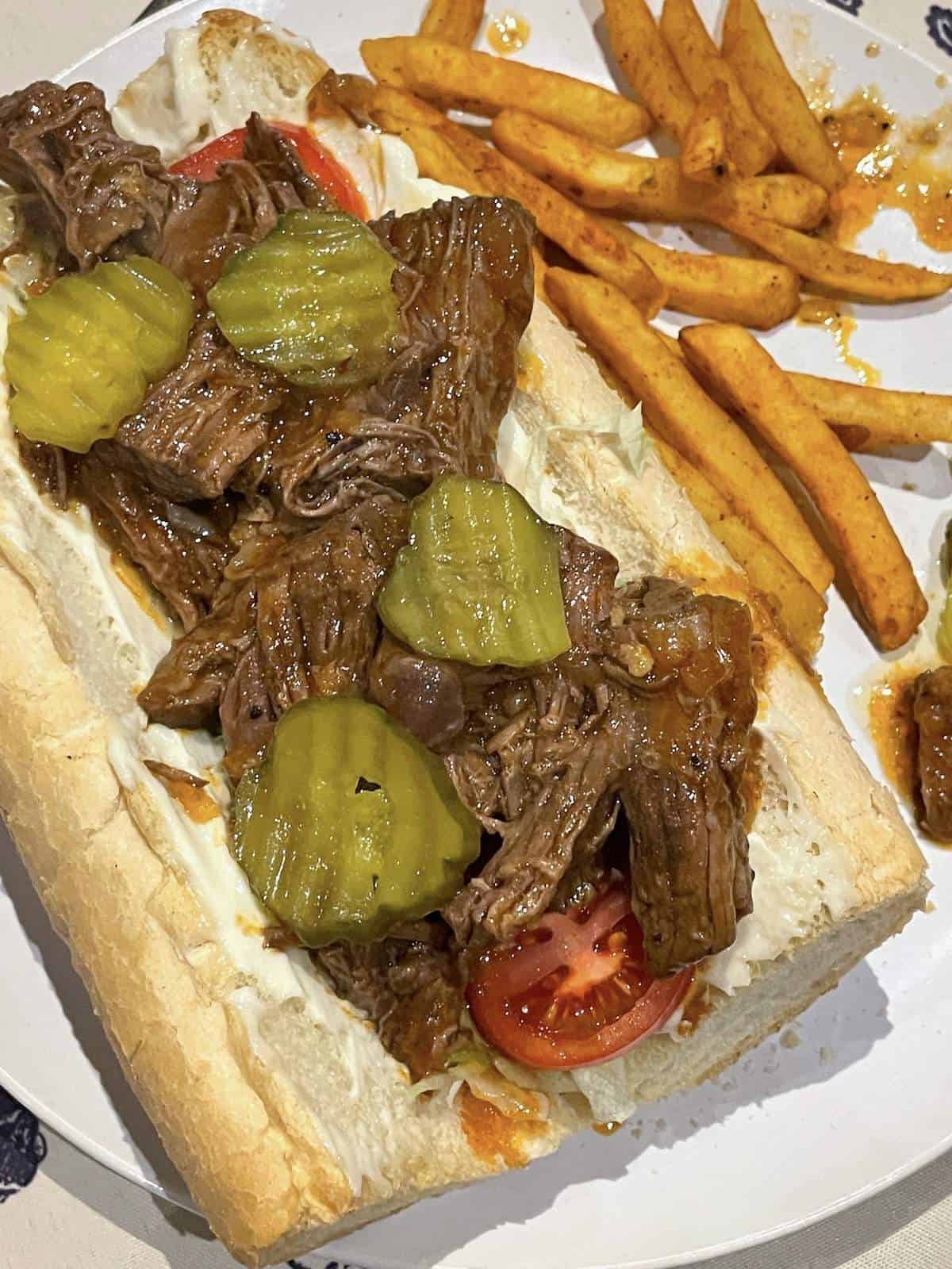 A roast beef debris po boy on French bread with lettuce, tomato, and pickles with fries on the side.