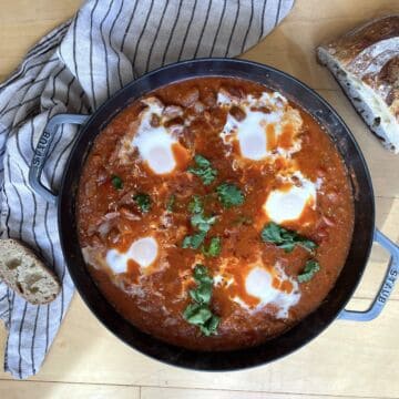 A pan of creole tomato shakshuka with 5 eggs and sourdough bread next to it.
