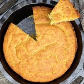 A cast iron pie pan with baked golden brown southern cornbread in it with a slice on top.