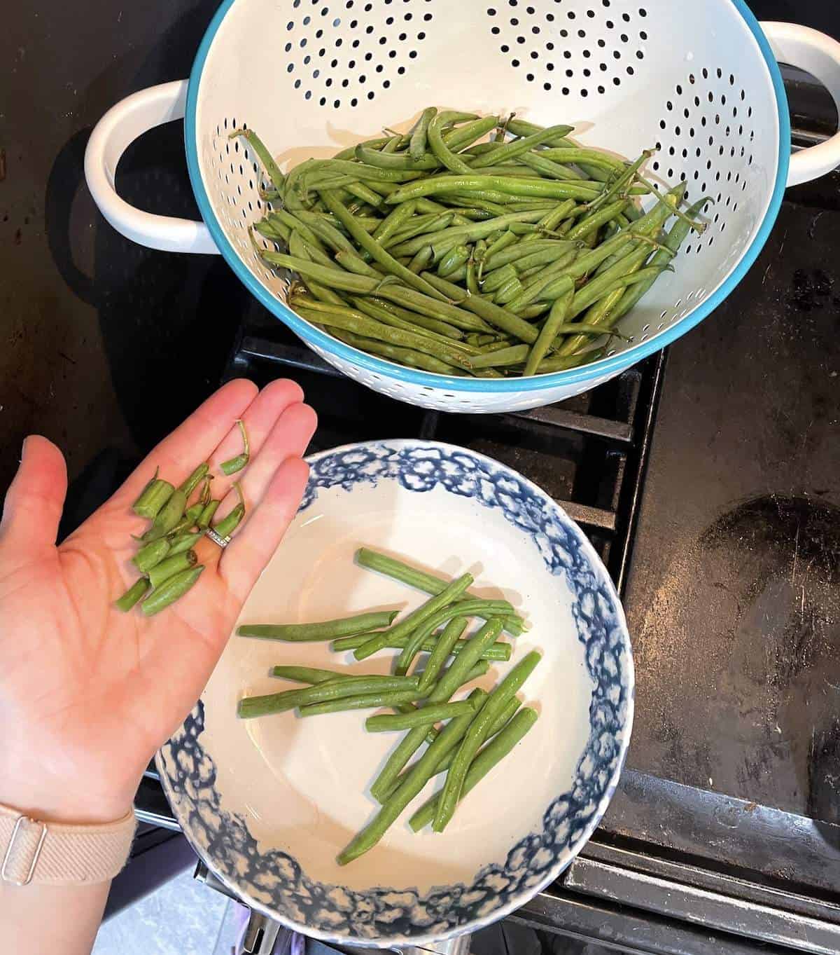 A colander of green beans and a bowl showing how to snap the ends off of fresh green beans.