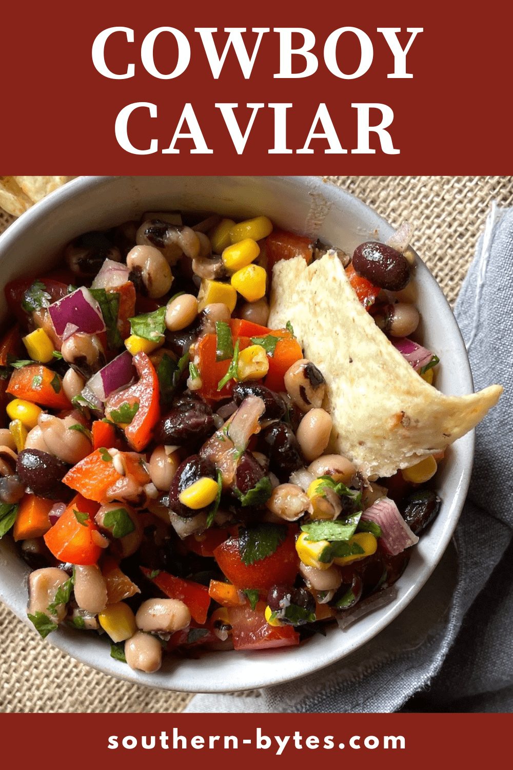 A pin image of a small bowl of cowboy caviar with black-eyed peas, black beans, corn, tomatoes, red onions, and cilantro with a vinegar based dressing and a side of tortilla chips.