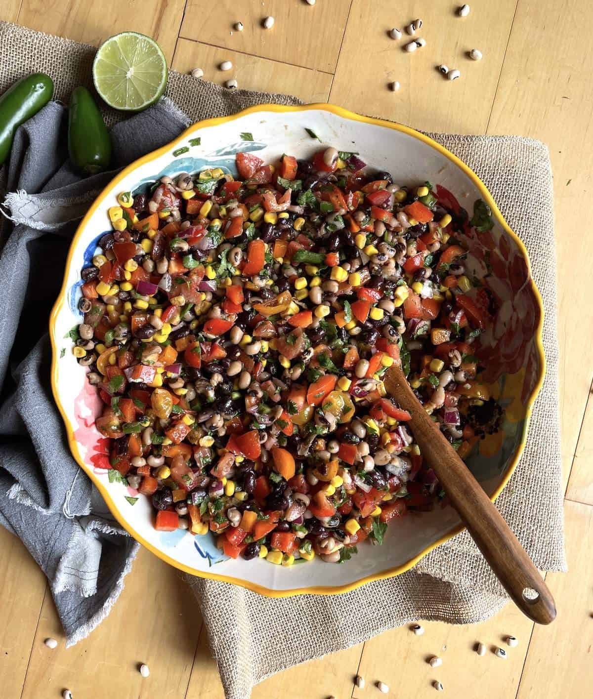 A bowl of redneck caviar with black-eyed peas, black beans, corn, tomatoes, cilantro, peppers, and a vinegar dressing with a wooden spoon.