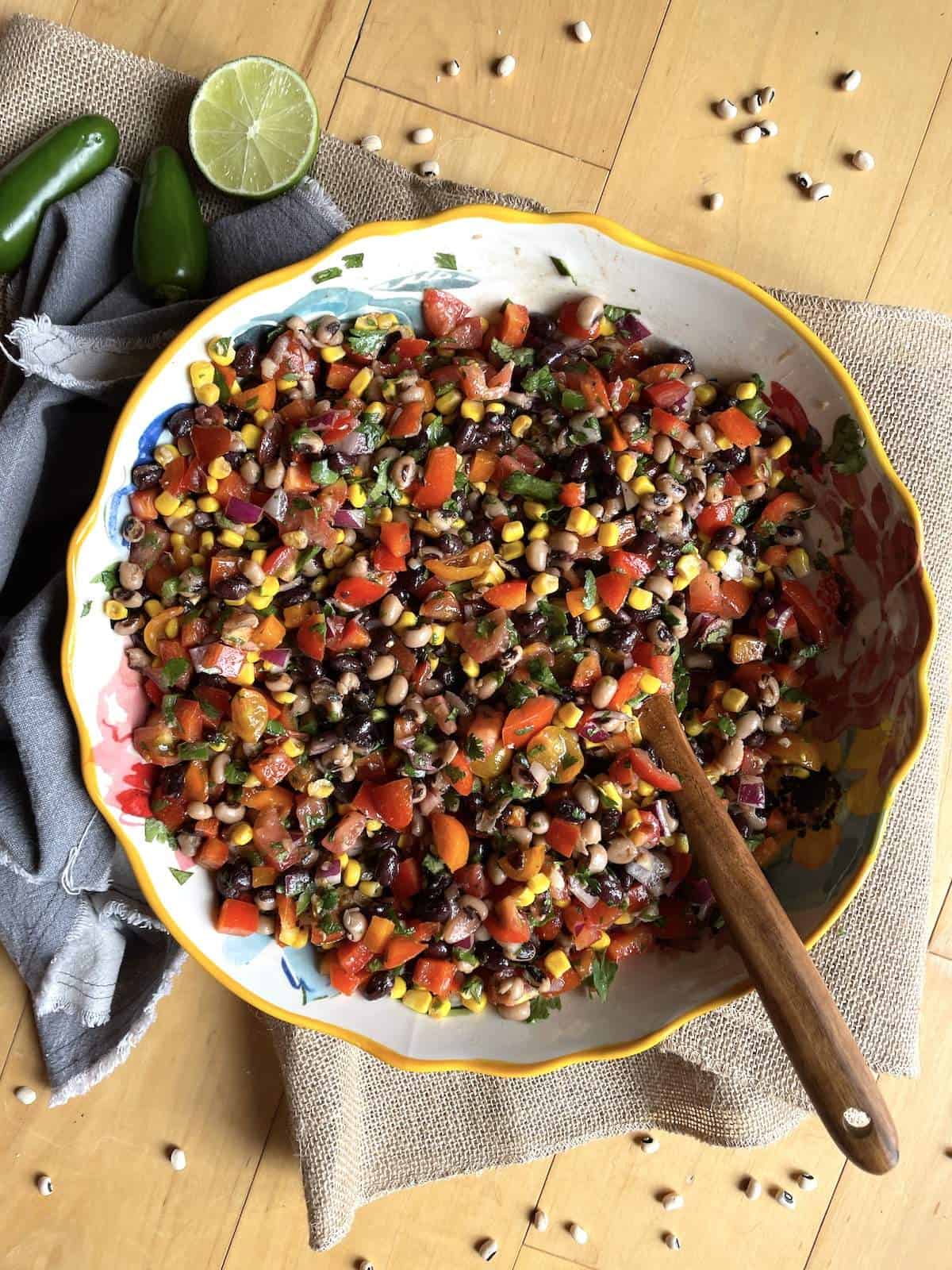 A yellow bowl of redneck caviar with black-eyed peas, black beans, corn, tomatoes, cilantro, peppers, and a vinegar dressing with a wooden spoon.