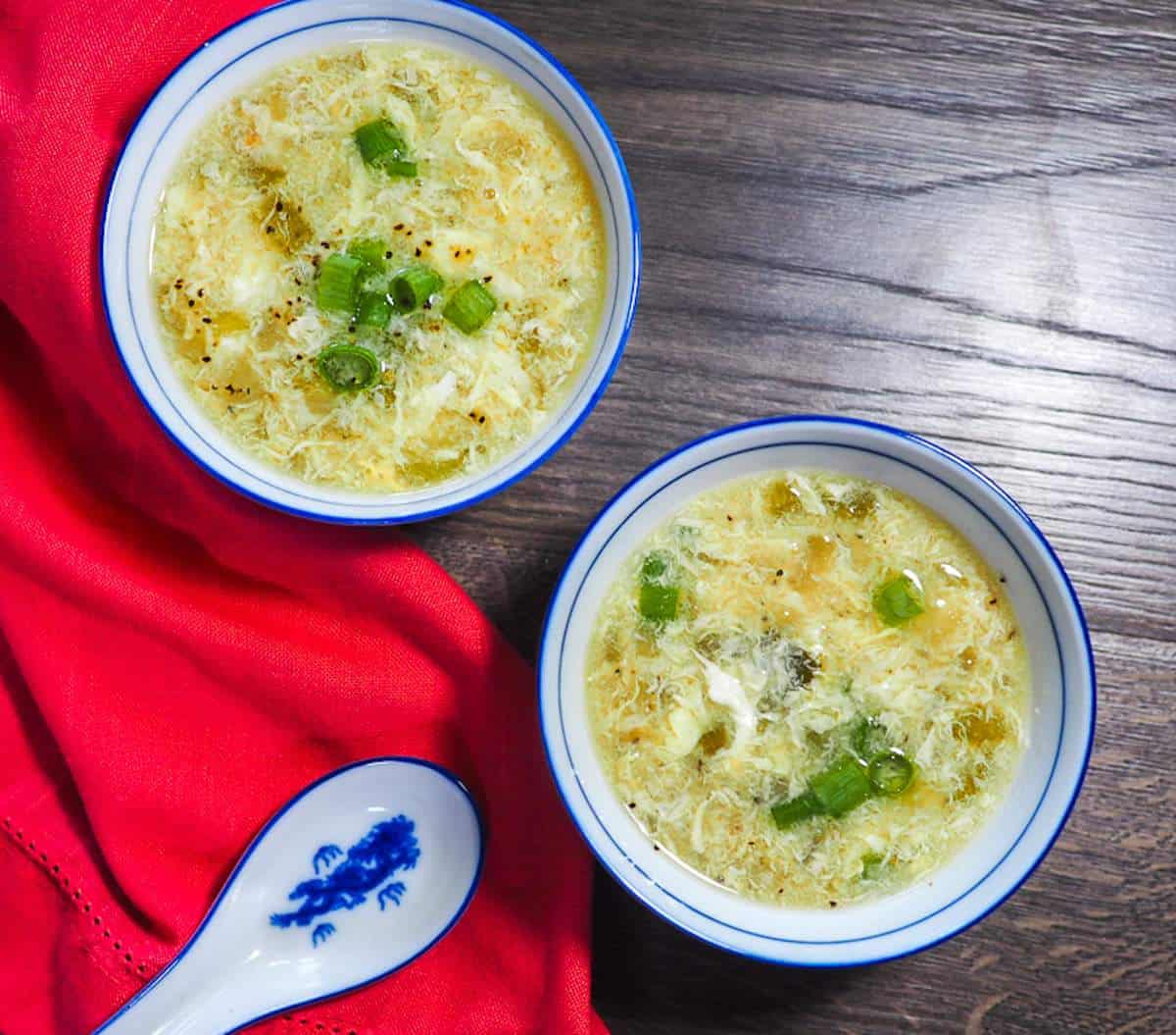 Two small blue and white bowls of homemade egg drop soup topped with sliced green onions.