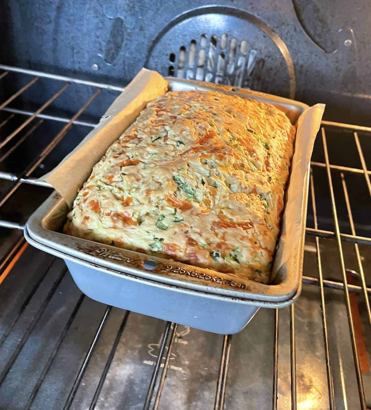 A loaf of savory zucchini bread with cheese and jalapeños baking in the oven in a parchment lined metal pan.