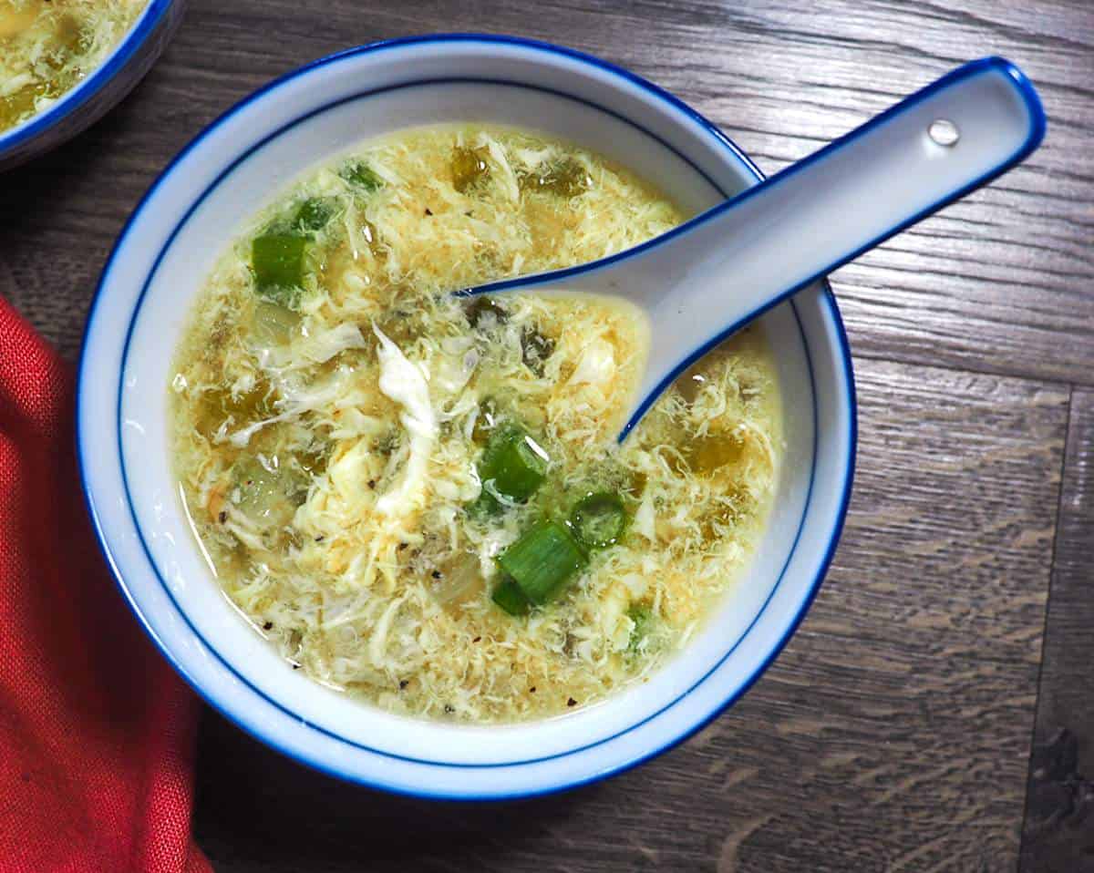 A small bowl of egg flower soup with a spoon - topped with sliced scallions.