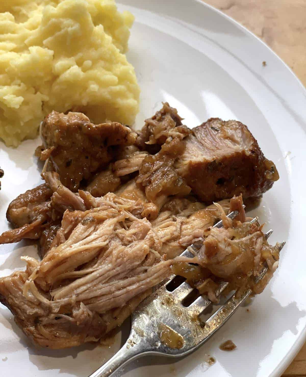 A white plate with shredded country style ribs and a scoop of buttermilk mashed potatoes with a fork.