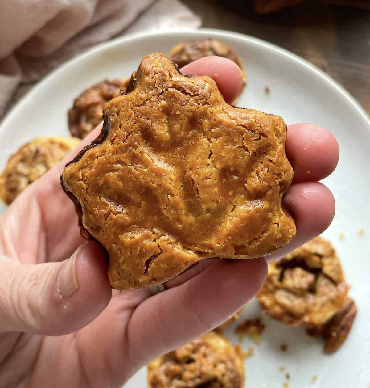A hand holding a maple leaf shaped pecan pie tart over a plate of tarts showing the back of the tart.