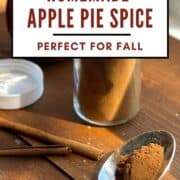 A pin image of an open jar of apple pie spice mix with some on a spoon and cinnamon sticks.