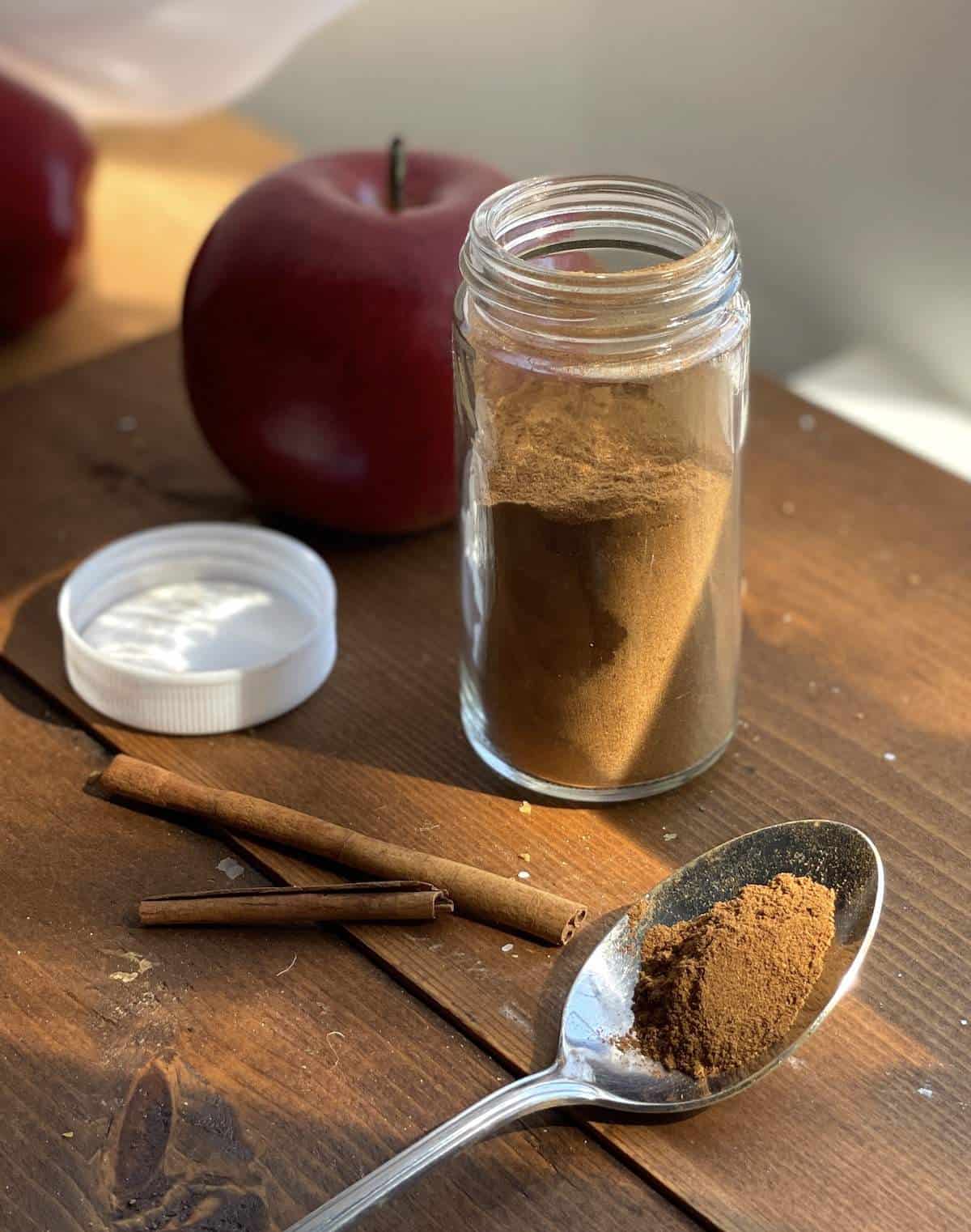 An open jar of apple pie spice mix with some on a spoon and cinnamon sticks.