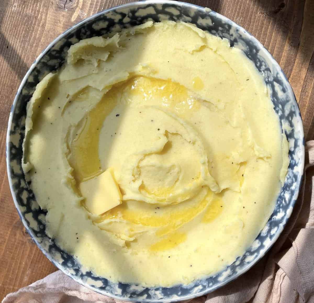A blue bowl of buttermilk mashed potatoes in the afternoon sunlight with melted butter on top.