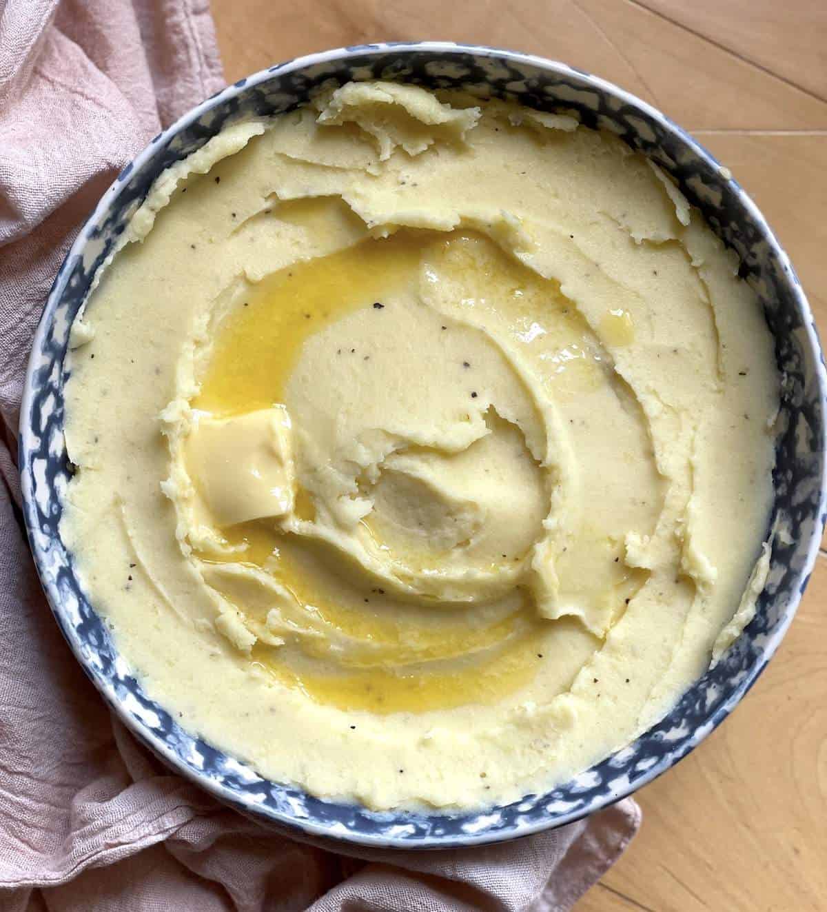 A blue bowl of buttermilk mashed potatoes with a pat of butter and some melted butter on top.