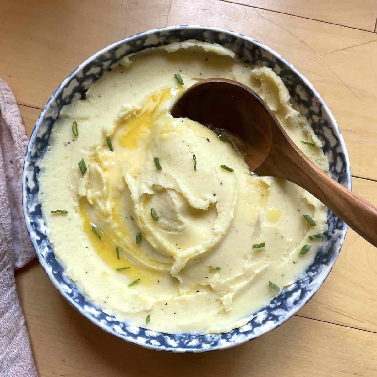 A blue bowl of buttermilk mashed potatoes with melted butter on top with a wooden serving spoon resting in it.