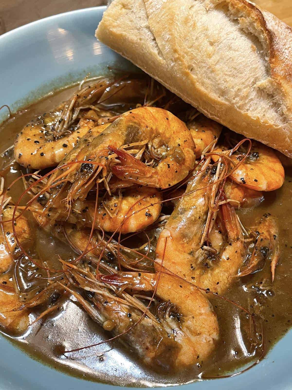 A blue bowl of New Orleans BBQ shrimp with a slice of bread.