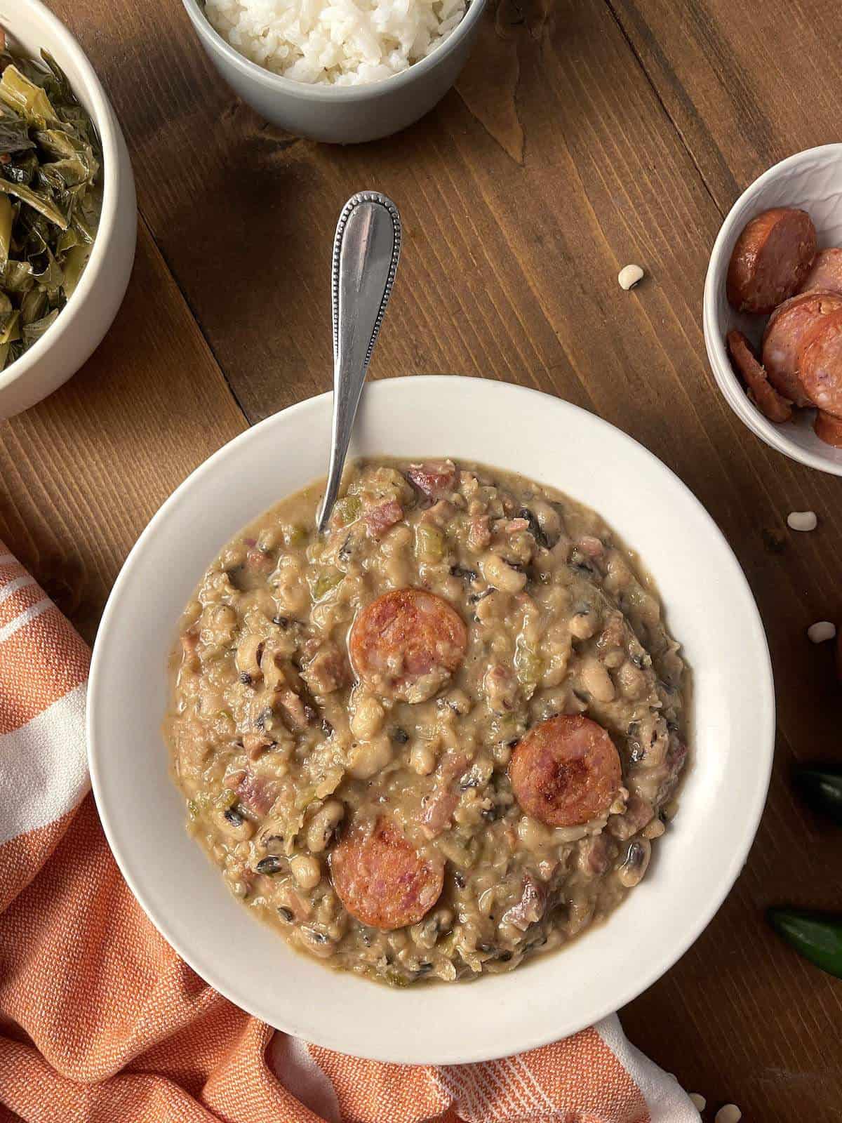 A large white bowl of hoppin' john black eyed peas with a small bowl of sausage, a bowl of rice, and some collard greens.