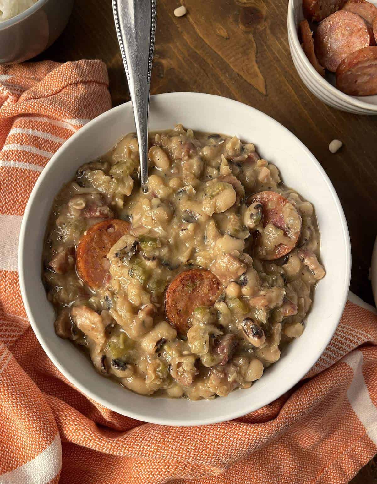 A white bowl of hoppin' john black eyed peas with a spoon, a small bowl of sausage, and an orange towel.