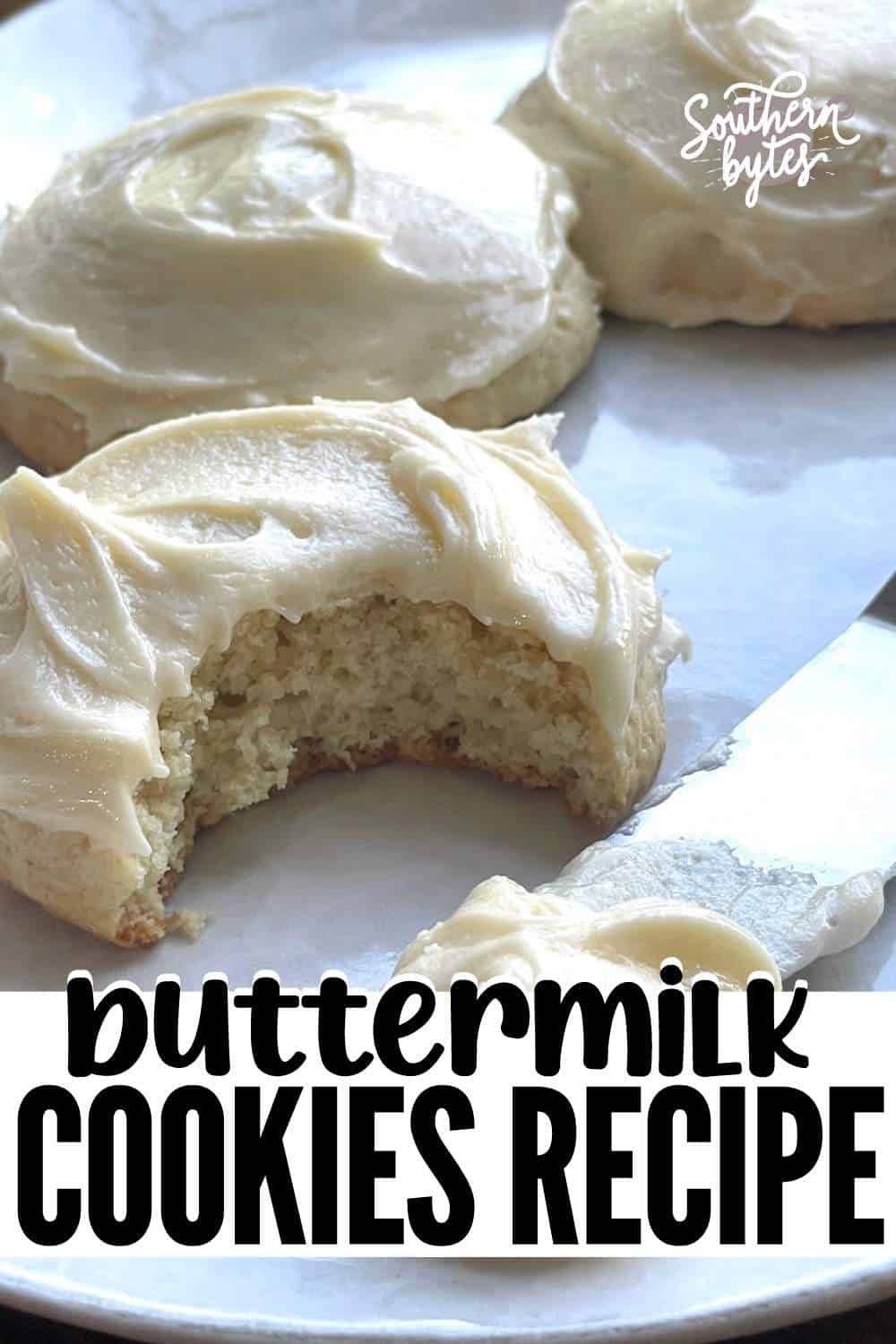 A pin image of a buttermilk cookie with thick frosting and a bite taken out of it with two cookies behind it.