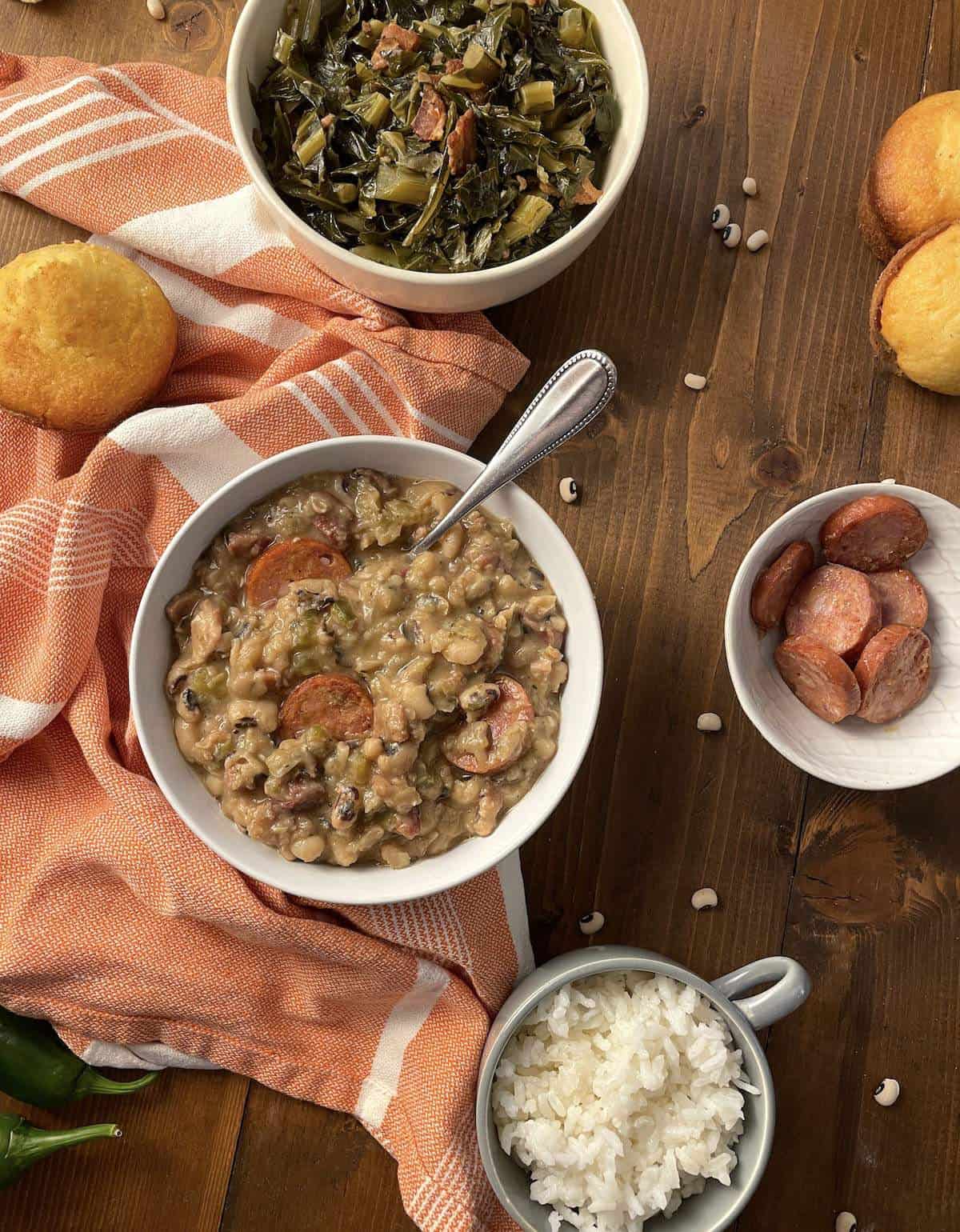 A bowl of cooked black eyed peas with collard greens, cornbread, rice, and sausage.
