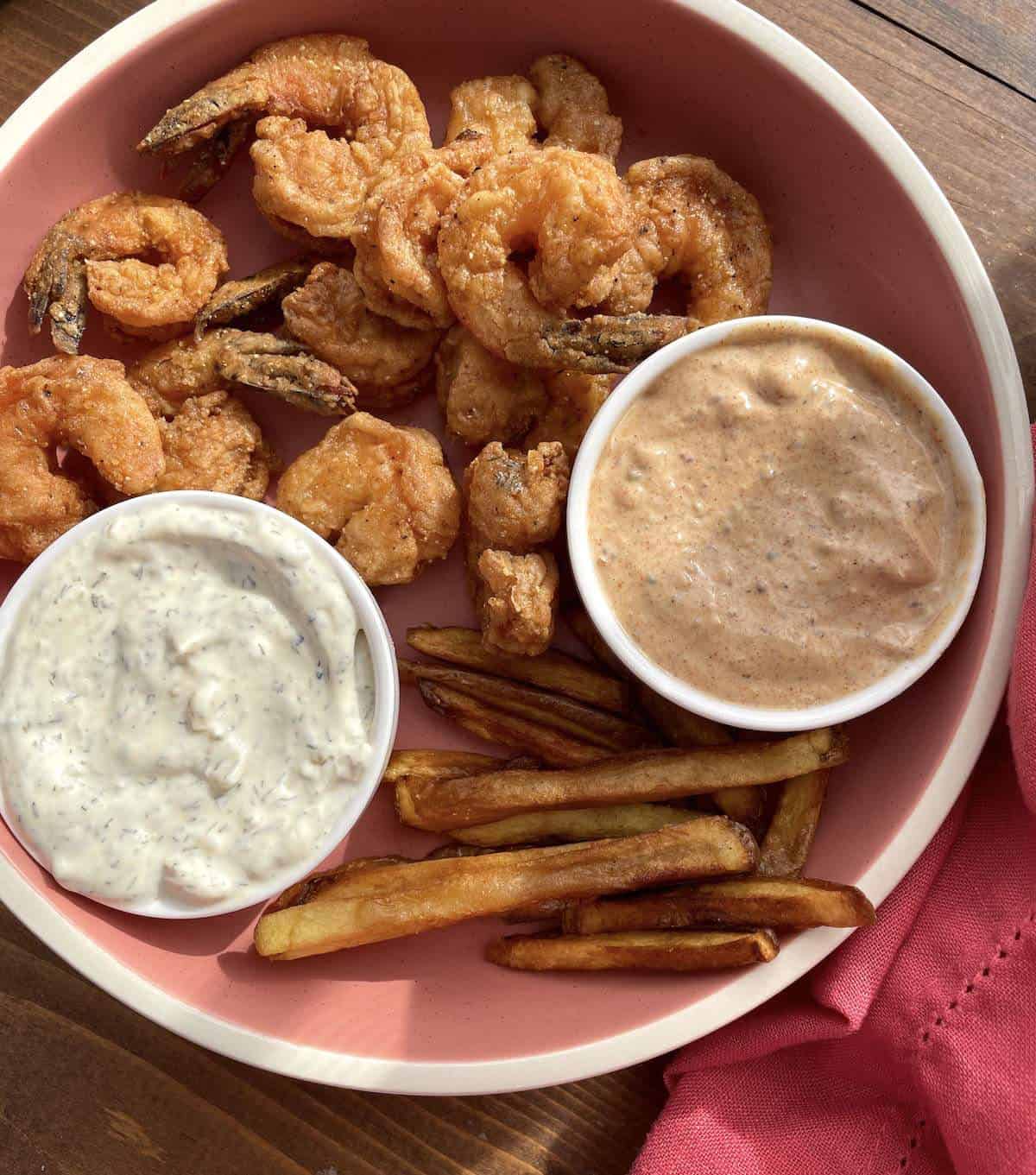 A pink bowl with french fries, fried shrimp, tartar sauce, and remoulade sauce.
