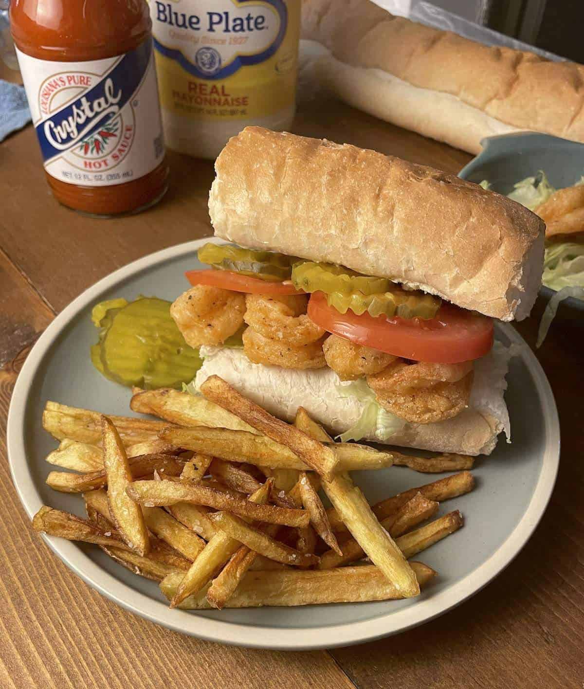 A fully dressed crispy fried shrimp po boy on a blue plate with french fries, and hot sauce.