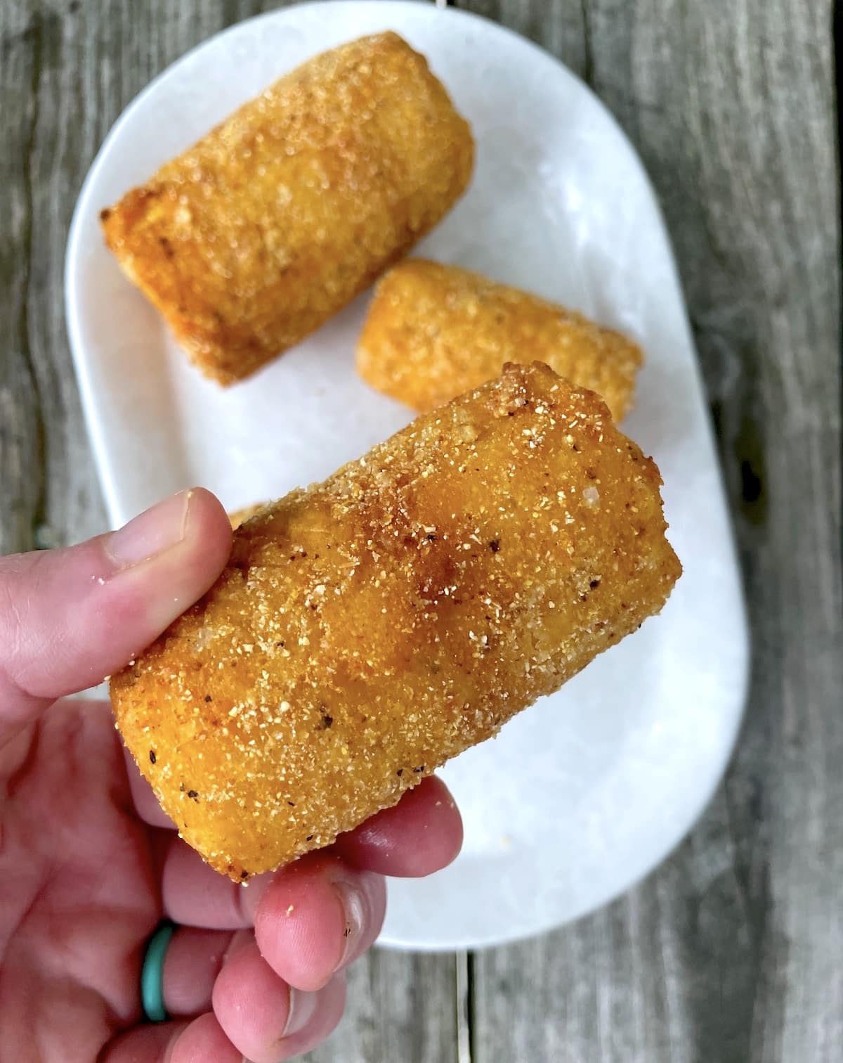 A hand holding a piece of fried corn on the cob over a white plate with two other pieces on it.