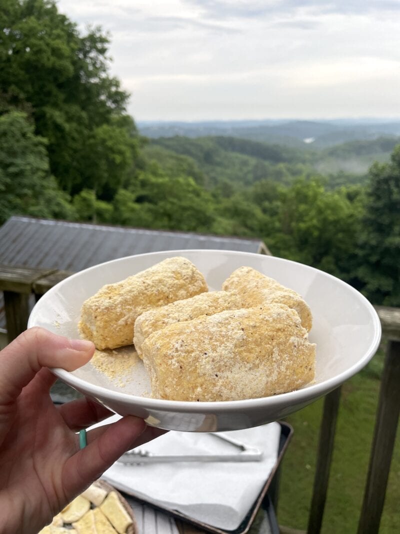 A bowl of cornmeal breaded corn on the cob, ready for frying, in front of a view of some mountains.