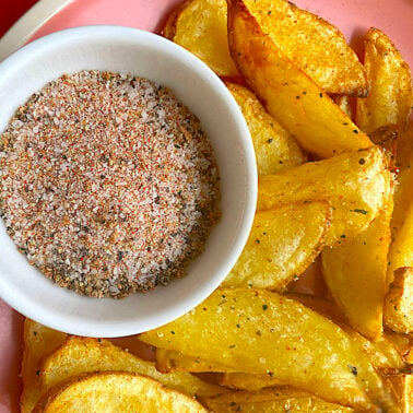 A bowl of Cajun sparkle fry seasoning next to seasoned potato wedges on a pink plate.