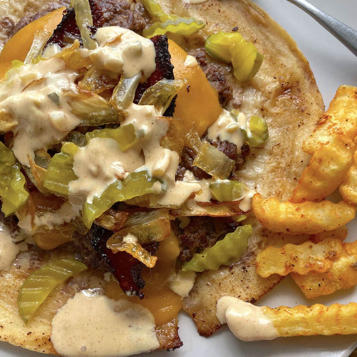 A smashed burger taco on a white plate topped with fried onions, pickles, cheddar cheese, and big mac burger sauce with fries on the side.