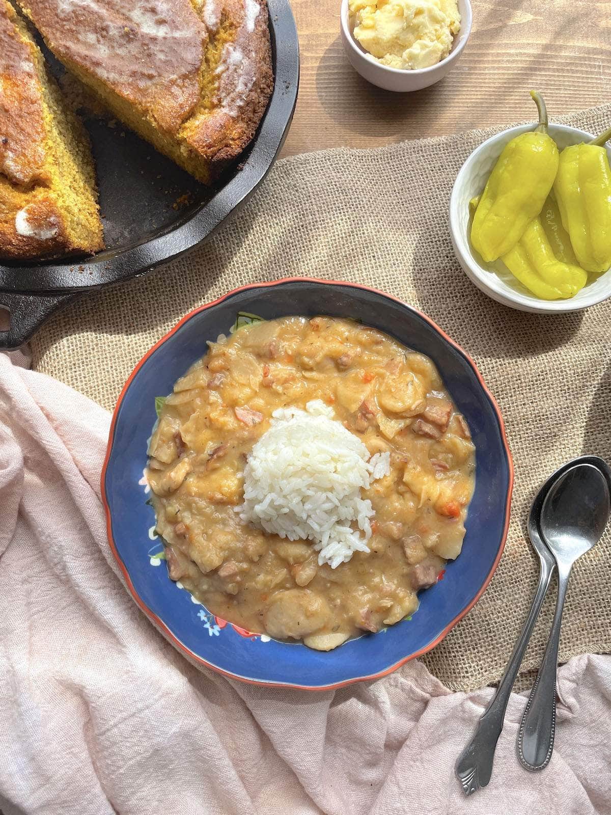 A blue bowl of butterbeans with a scoop of rice on top, pepperoncini peppers, and honey butter and sweet potato cornbread on the side.