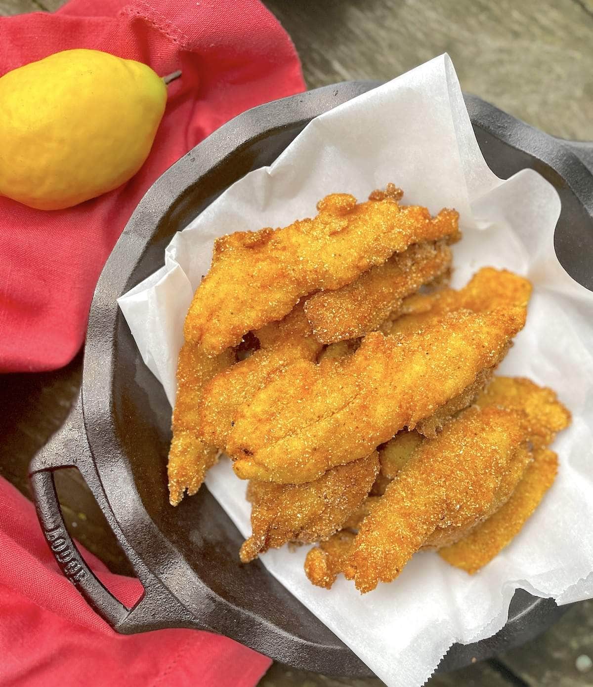 A cast iron pan filled with cornmeal crusted fried fish next to a lemon.