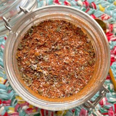 A jar of homemade creole seasoning on a colorful mat with a gold spoon on the side.