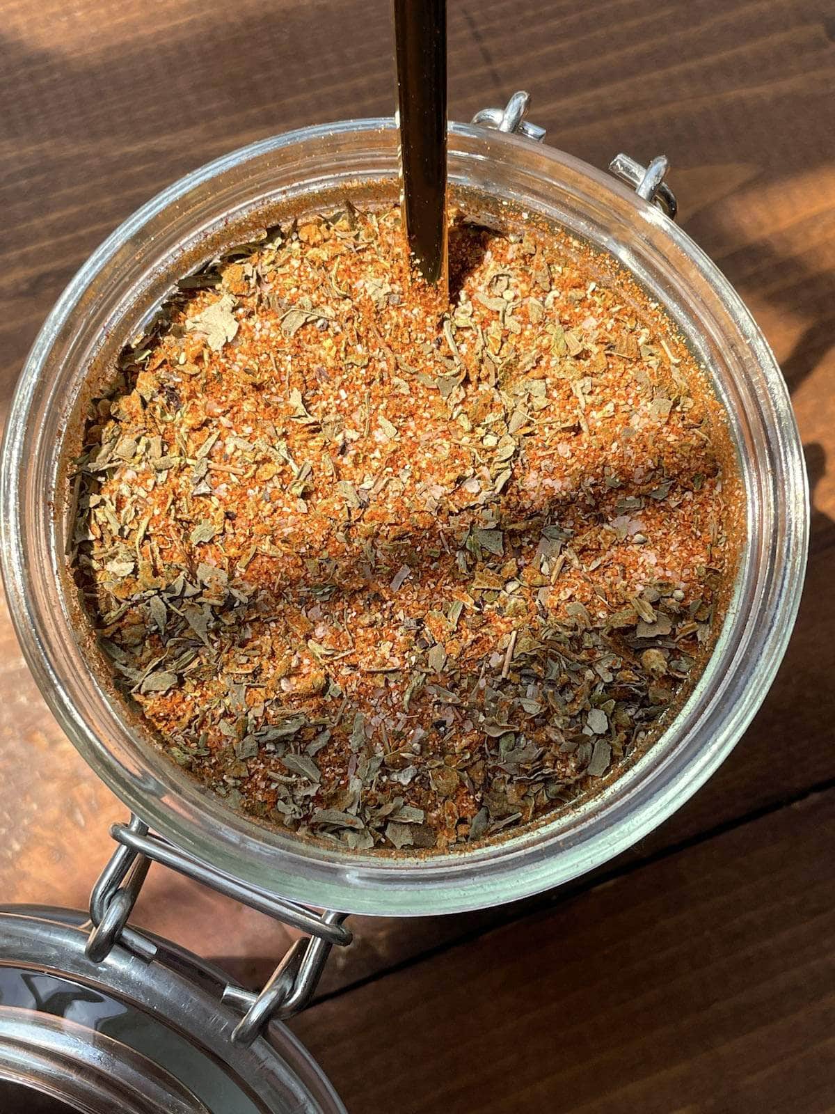 A jar of homemade creole seasoning on a brown background with a gold spoon in the jar.