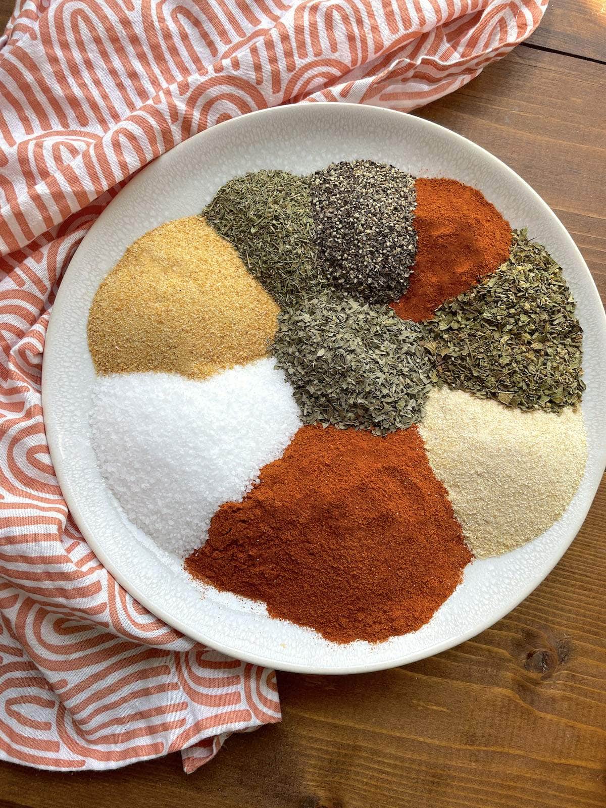 A white plate with the spices in Creole seasoning laid out on it next to a pink and white tea towel.