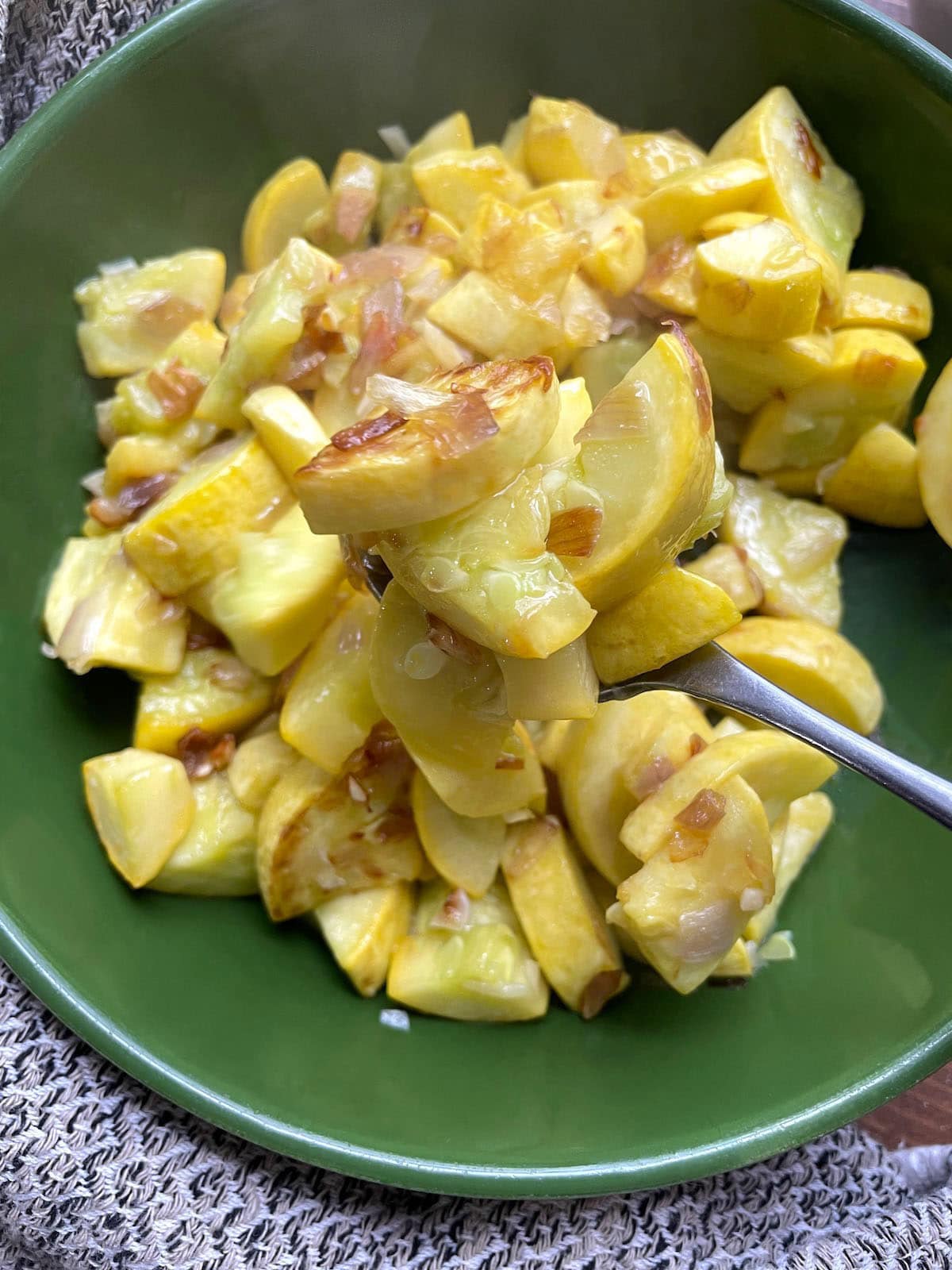 A green bowl filled with sauteed yellow squash and onions and some being scooped on a spoon.