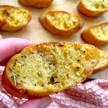 A hand holding buttered garlic toast over several slices on a wooden board.