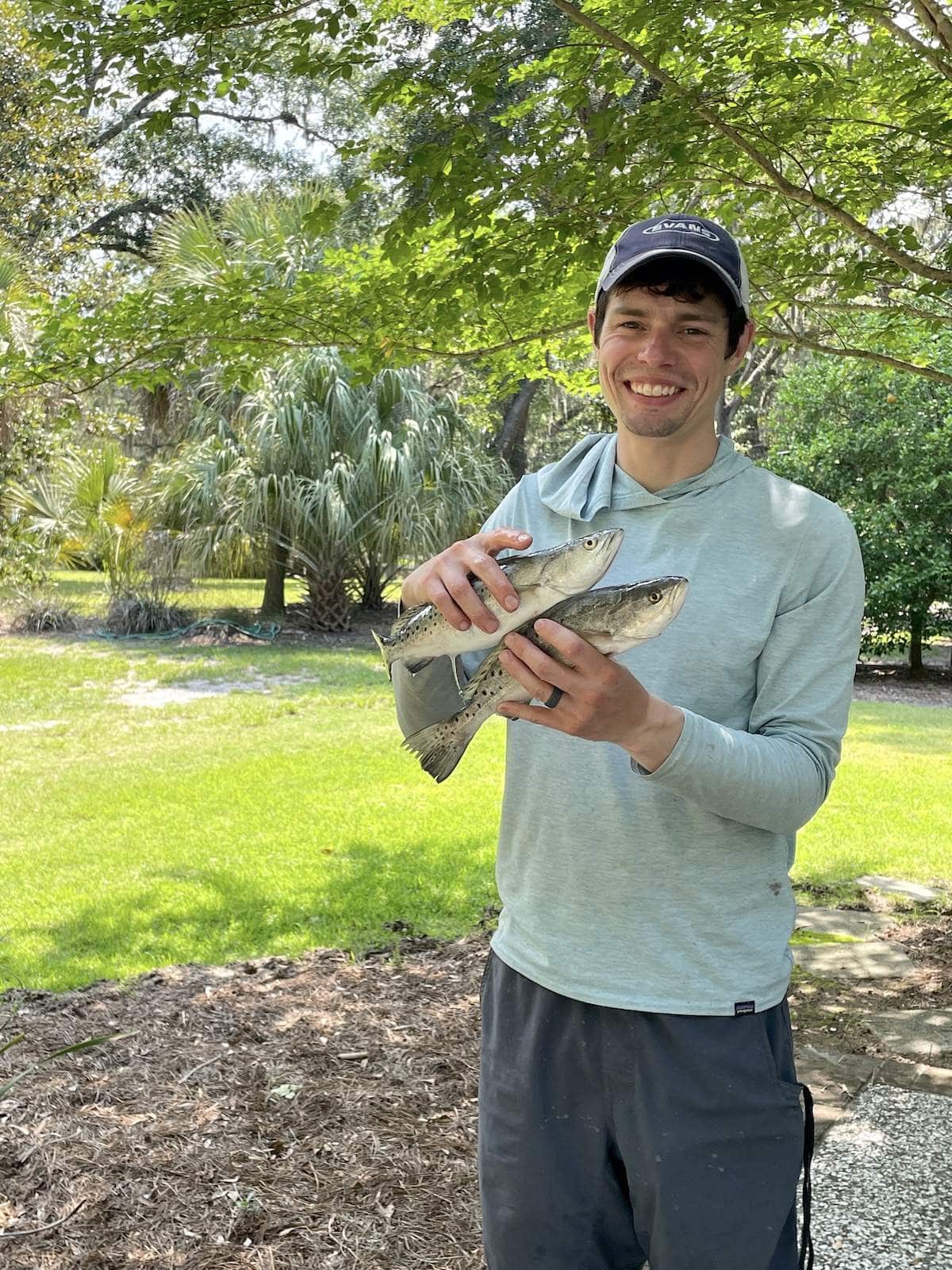 A man in a blue shirt wearing a hat holding two speckled trout.