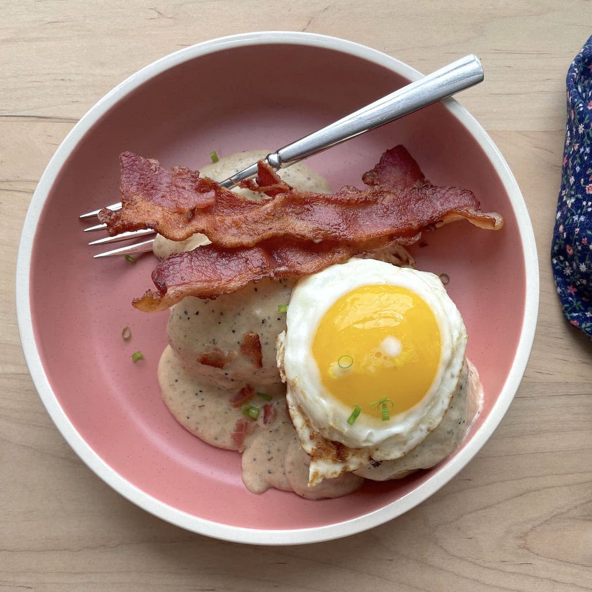 Two biscuits with two strips of bacon topped with bacon gravy in a pink bowl topped with a fried egg.