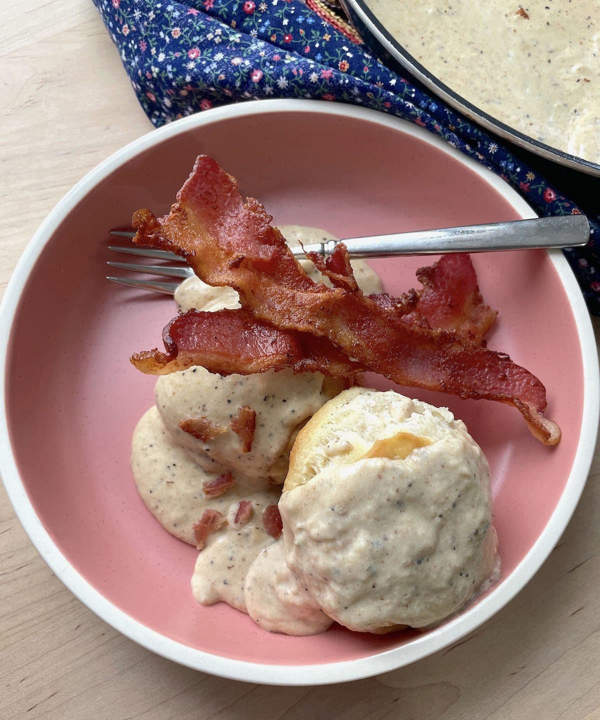 Two biscuits with two strips of bacon topped with bacon gravy in a pink bowl with a fork.
