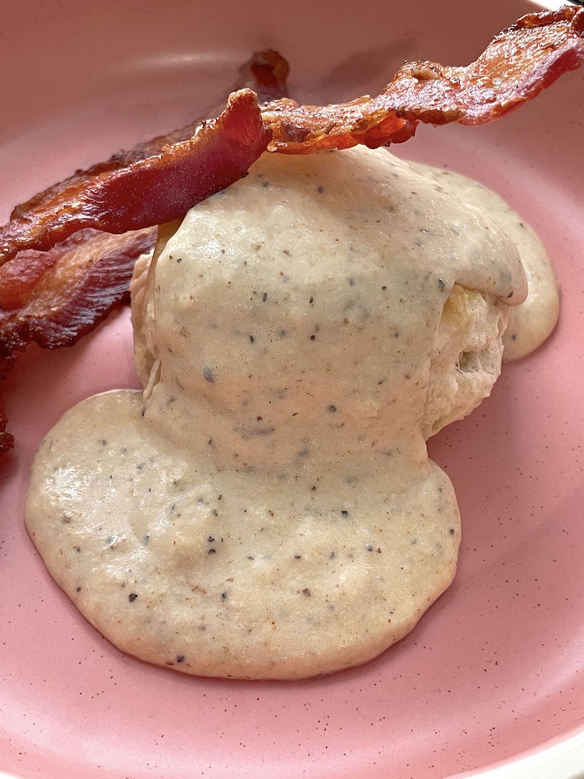 Two biscuits with two strips of bacon smothered with bacon gravy in a pink bowl.