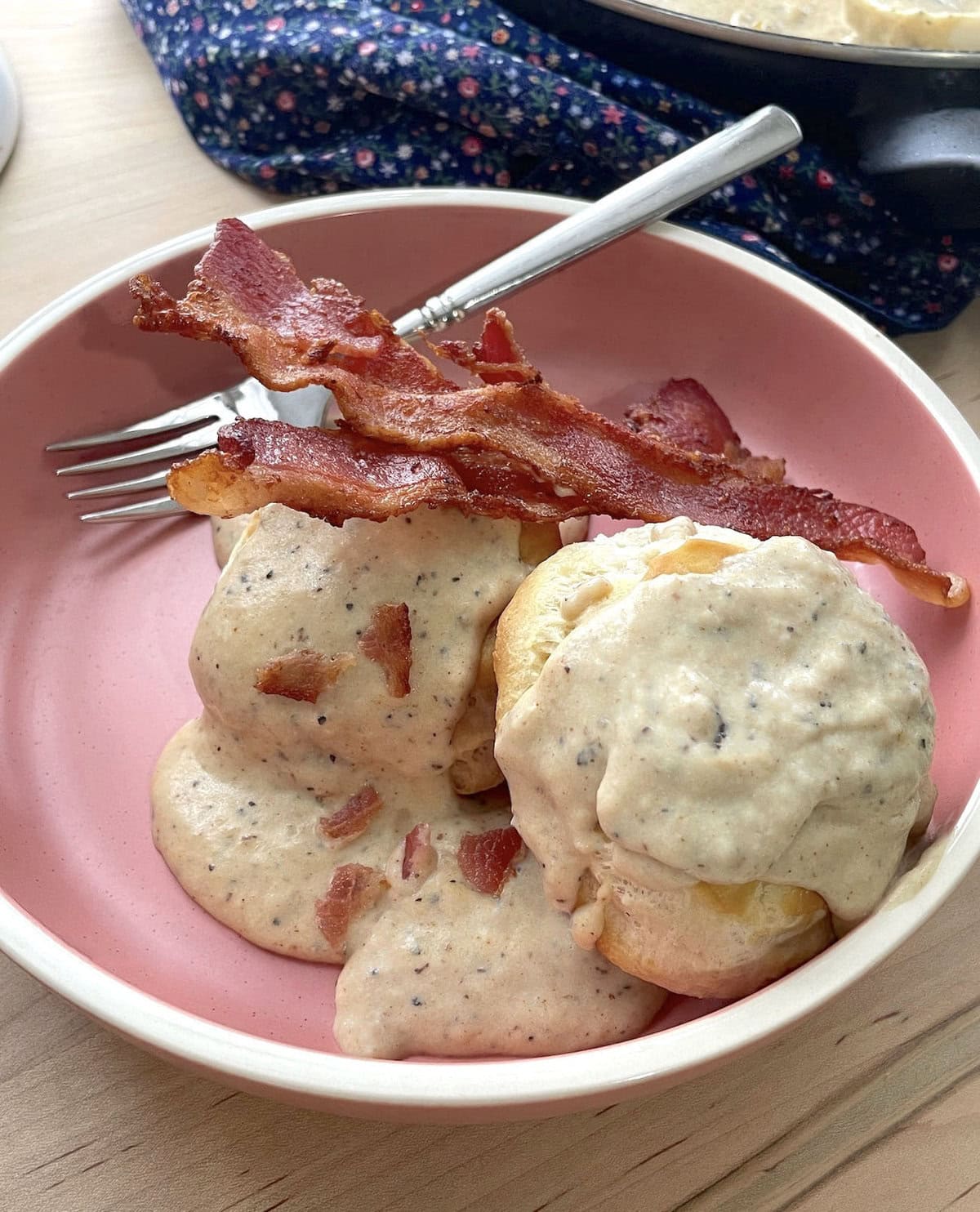 Two biscuits with two strips of bacon topped with bacon gravy in a pink bowl with a fork.