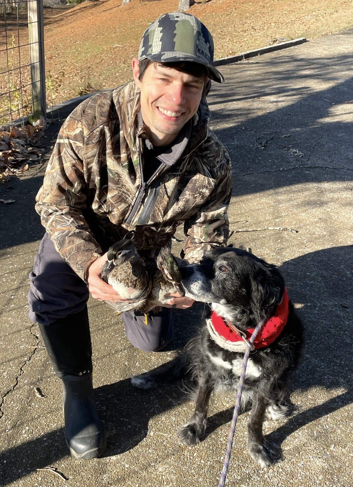 A man in camouflage holding two ducks and a black dog sniffing them.