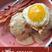 A pin image of two biscuits with two strips of bacon topped with bacon gravy in a pink bowl topped with a fried egg.
