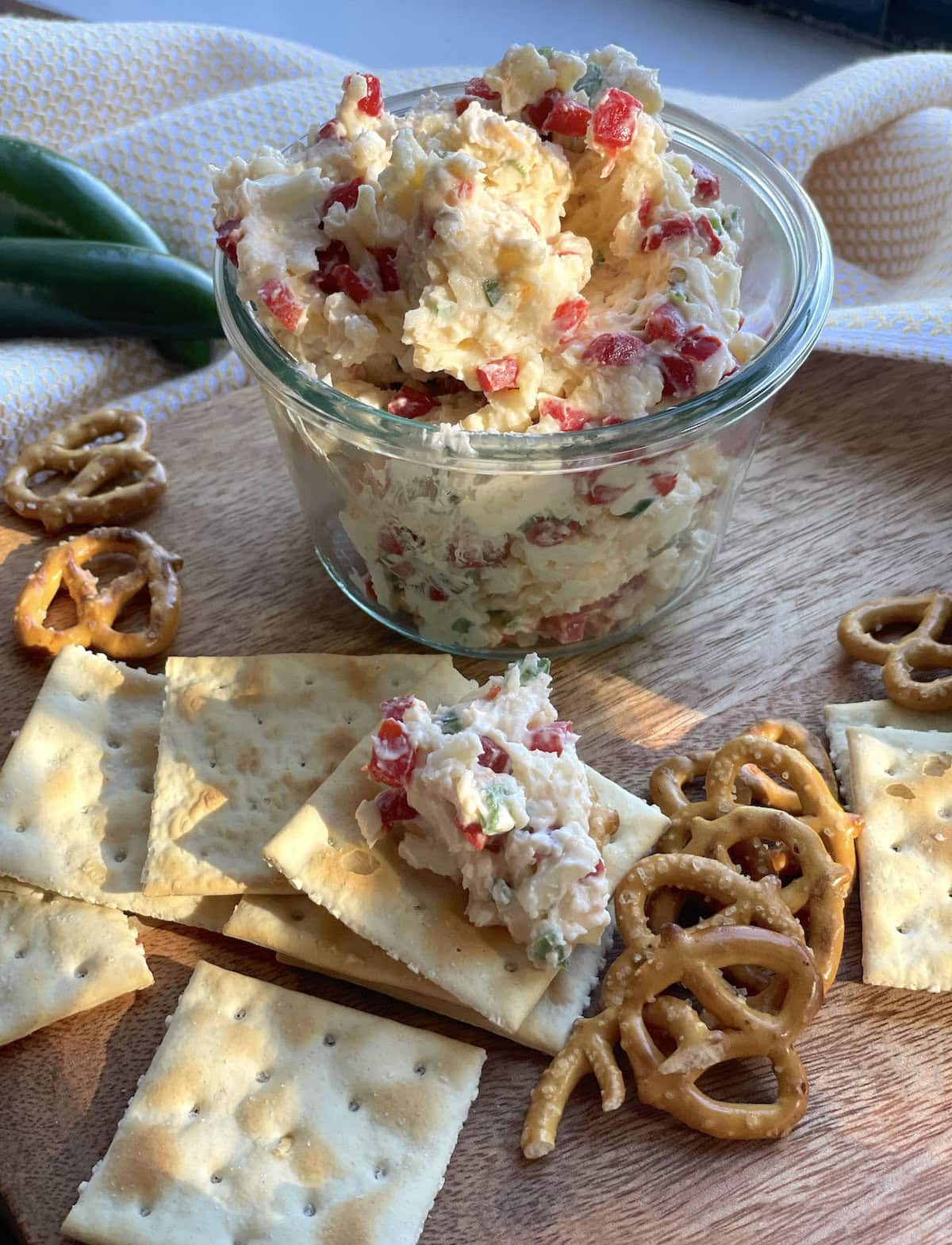 A jar of pimento cheese with jalapeños surrounded by saltines and pretzels.