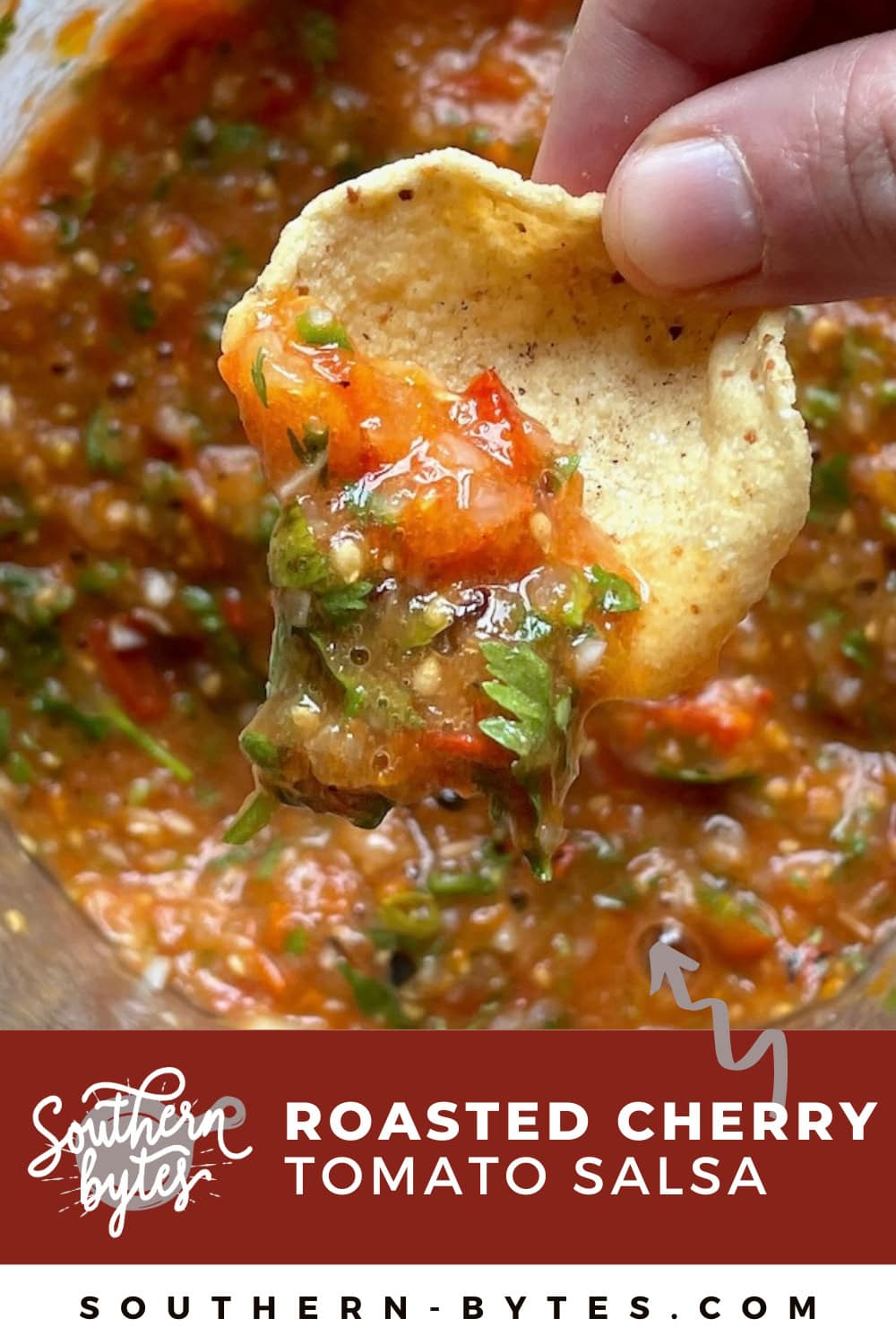 A pin image of a hand scooping some fire-roasted cherry tomato salsa up with a chip.