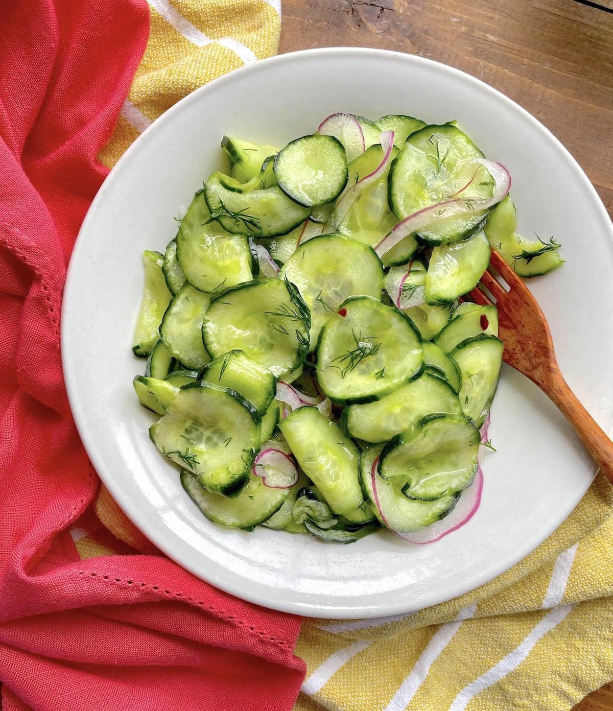 A white bowl with pickled cucumber salad - thinly sliced cucumbers, red onions, and lots of dill with wooden utensils and pink and yellow towels.