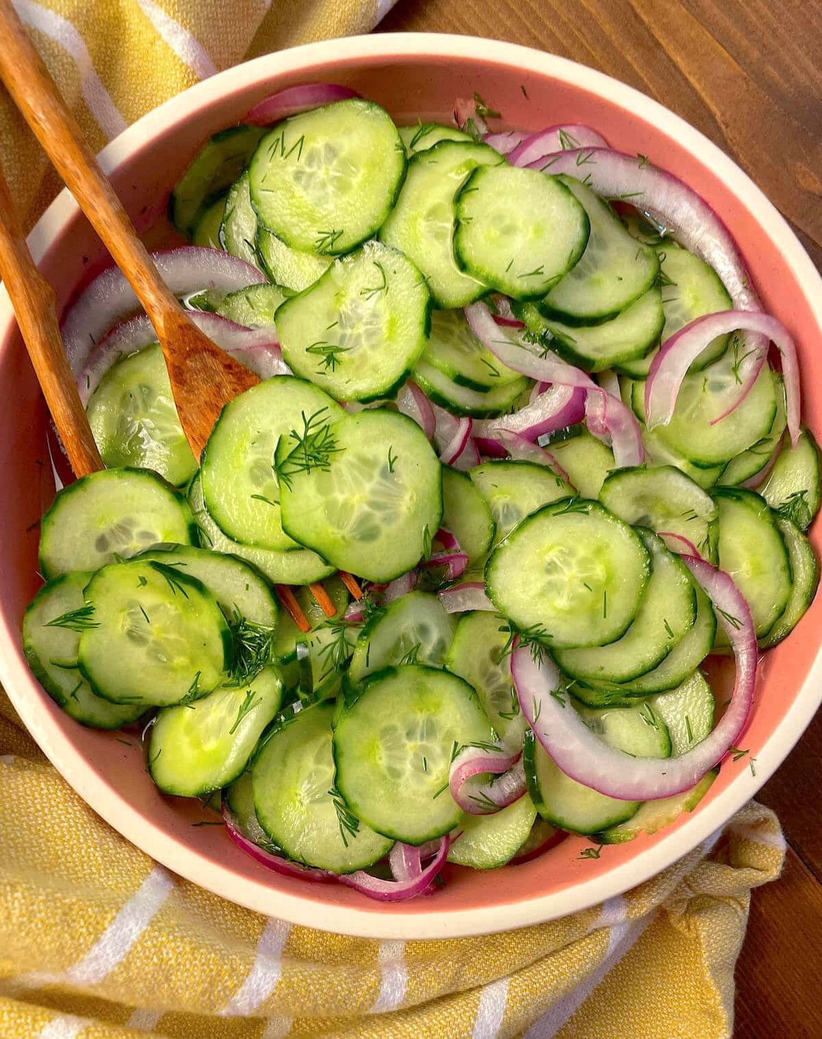 A pink bowl with pickled cucumber salad -thinly sliced cucumbers, red onions, and lots of dill with wooden utensils and a yellow towel.