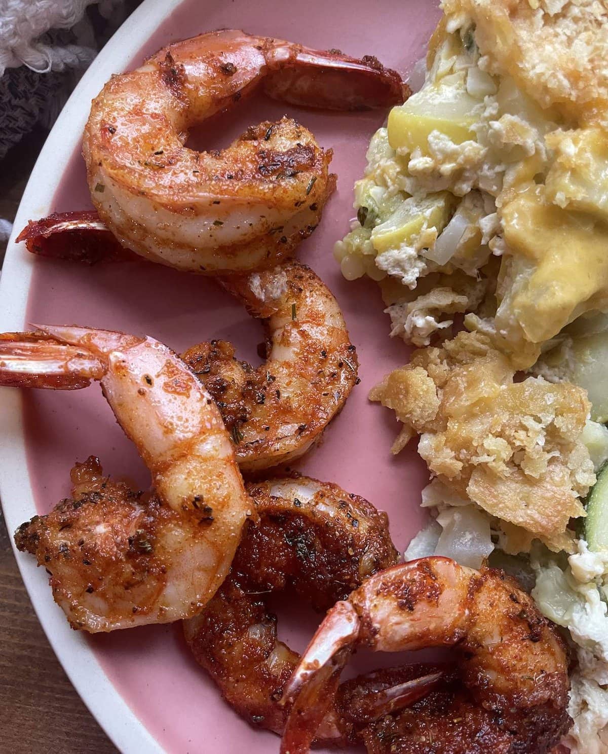 Blackened shrimp with tails on laid out on a pink plate with squash casserole.