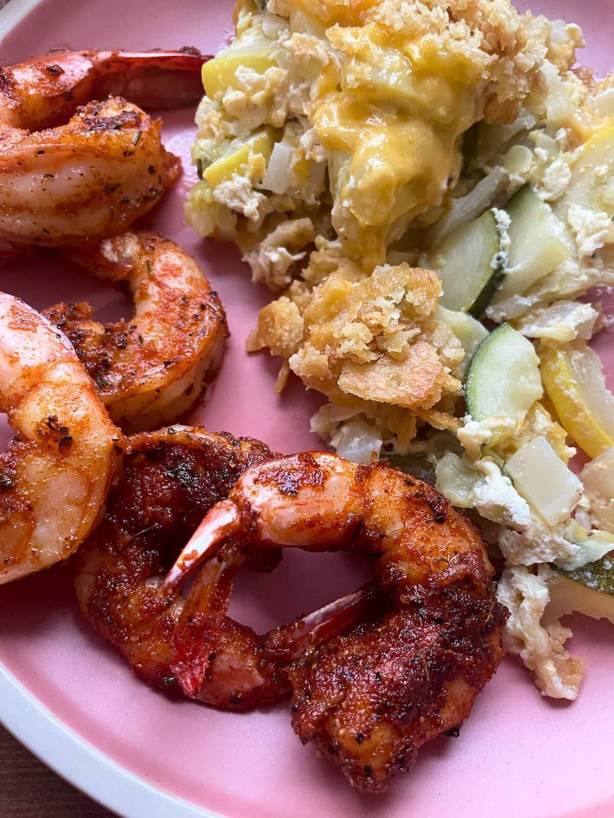 Blackened shrimp with tails on sitting on a pink plate with squash casserole.