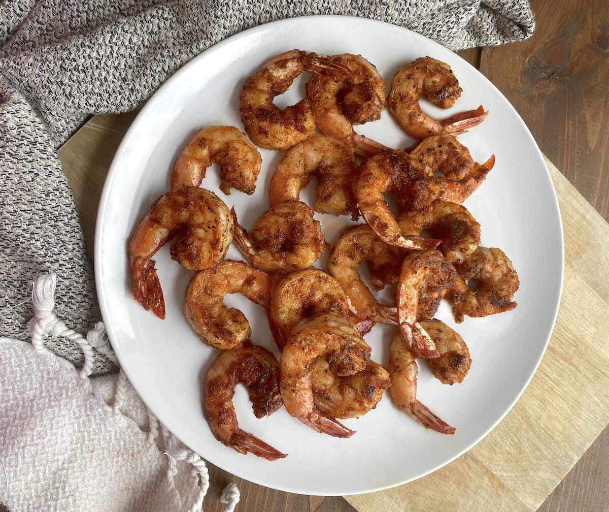 A white plate of blackened shrimp with tails on them sitting on a wood board with a tea towel next to it.