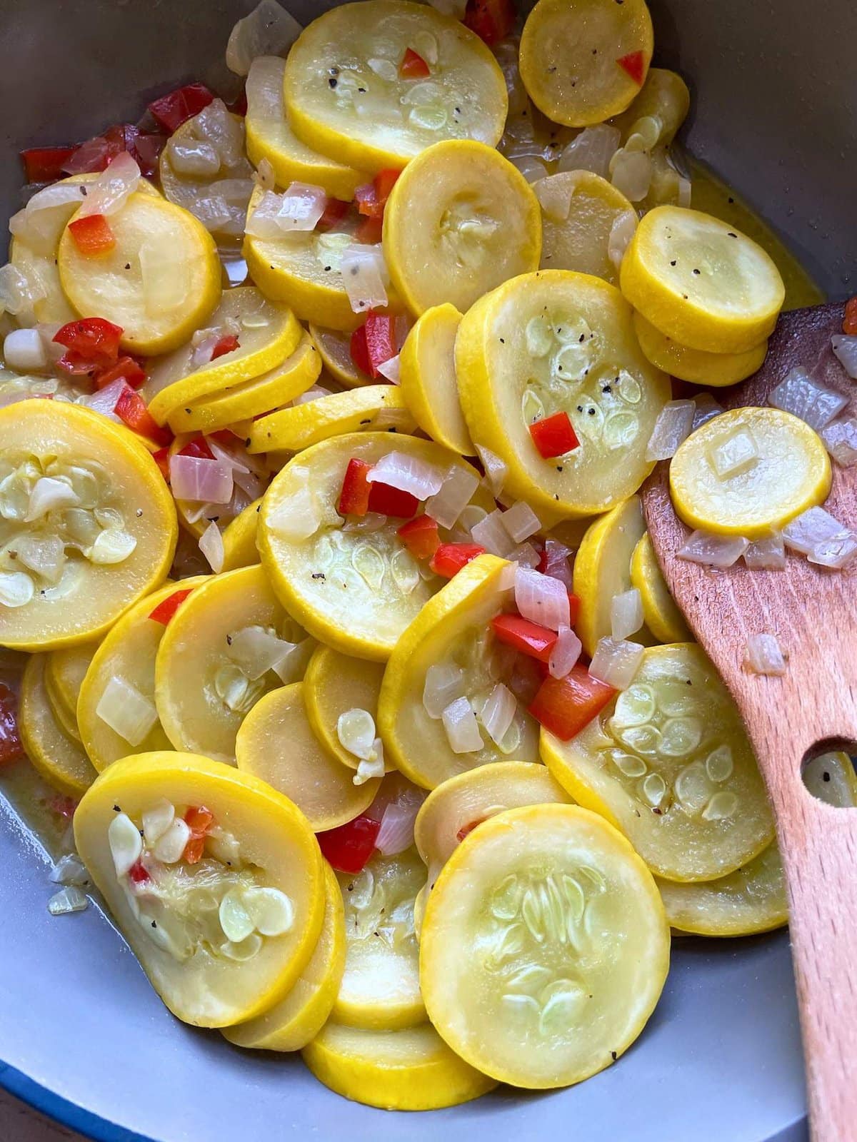 A blue and gray frying pan with sautéed squash, red bell peppers, and onions in it with a wooden spoon.